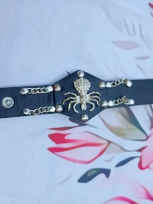 Svart armband med spindeldekoration - Unikt svart armband med silverfärgade kedjor och nitar samt en stor spindeldekoration i metall framtill. Bältet har tryckknappsstängning och detaljer som ger en edgy look. Perfekt för dig som vill sticka ut med accessoarerna.