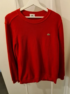 Röd sweatshirt från Lacoste - Klassisk röd sweatshirt från Lacoste med rund halsringning och det ikoniska gröna krokodilmärket broderat på bröstet. Tröjan har ribbade muddar vid ärmslut och nederkant. Perfekt för en clean och sportig look.