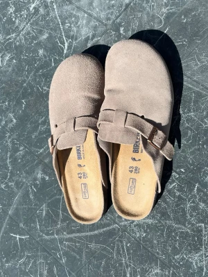 Beige mockatofflor från Birkenstock - Säljer ett par klassiska beige tofflor från Birkenstock i mjuk mocka med justerbart spänne och ergonomisk fotbädd. Perfekta för dig som vill ha både stil och komfort. Passar till dig som gillar en avslappnad look.