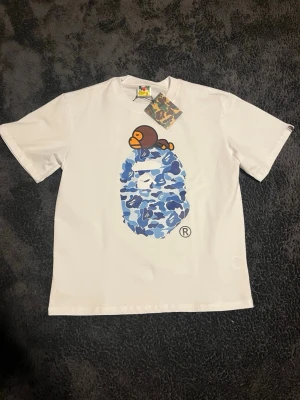 Vit BAPE t-shirt med blått camo-tryck - Vit bape tröja med blått camo tryck på framsidan. Tröjan är helt oanvänd med lapp kvar. Hör av er vid frågor 