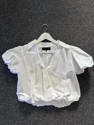 Vit puffärmad cropped blus - Trendig vit blus med puffiga korta ärmar och bred krage. Modellen är croppad med resår i nederkanten och har en djup V-ringning framtill. Perfekt för en stilren och modern look. Aldrig använd, endast provad. 