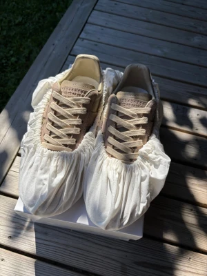 Beige maison margiela gats - Snygga sneakers i beige och ljusbrun färg med ovandel i mocka och släta läderpaneler. Skorna har vita snören och en klassisk gummisula. Perfekta för dig som gillar stilrena och neutrala färger.