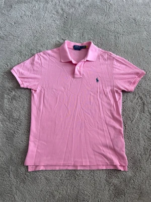 Rosa pikétröja från Polo Ralph Lauren - Klassisk rosa pikétröja från Polo Ralph Lauren med blå logga broderad på bröstet. Tröjan har korta ärmar, krage och två knappar framtill. Perfekt till sommaren och tillverkad i mjuk bomull för en bekväm känsla.