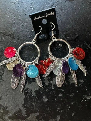 Stora boho örhängen med fjädrar - Unika örhängen i boho-stil med stora silverfärgade ringar, hängande metallfjädrar och färgglada runda pärlor i rött, blått, lila, vitt och gult. Perfekta för dig som gillar statement-smycken och vill sticka ut med färg och form.