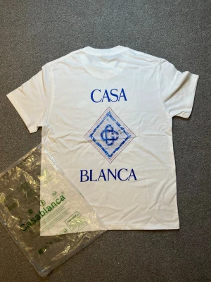 Vit t-shirt med blått tryck Casablanca - Vit t-shirt från Casablanca med stort blått tryck på ryggen där det står 'CASA BLANCA' och ett grafiskt mönster i mitten. Klassisk passform och rund hals. Perfekt för dig som gillar stilrena plagg med en twist.
