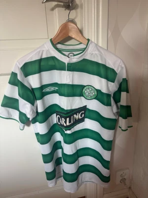 Grönvit Celtic matchtröja Umbro - Säljer en klassisk Celtic FC matchtröja från Umbro med gröna och vita horisontella ränder. Tröjan har klubbmärke på bröstet, Carling-logga på magen och rund hals. Tröjan är från Celtics 100 års jubileum som förening.