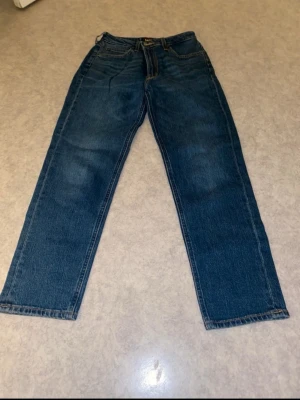 Mörkblå raka jeans från Lee - Säljer ett par klassiska mörkblå jeans från Lee med rak passform och fem fickor. Jeansen har hög midja och tydliga sömmar i kontrastfärg. Tillverkade i slitstarkt denimtyg, perfekta för en avslappnad och tidlös stil.