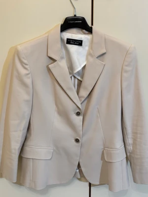 Beige kavaj från Caractère - Stilren beige kavaj från Caractère med två knappar framtill och klassisk krage. Kavajen har två fickor med lock och är figursydd för en snygg siluett. Materialet ser ut att vara slätt och lätt, perfekt för en dressad look.