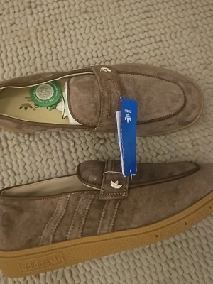 Bruna mocka loafers från Adidas - Snygga loafers i brun mocka från Adidas med klassiska detaljer och gummisula. Skorna har dekorativa sömmar, diskret Adidas-logga på sidan och ovandelen samt en stilren siluett. Perfekta för dig som gillar retrovibbar och streetstyle.