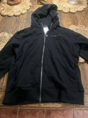 Svart hoodie från Sail Racing - Svart hoodie från Sail Racing med dragkedja framtill och logga på bröstet. Klassisk modell med huva och ribbade muddar. Perfekt för en avslappnad och sportig stil. Aldrig använt då en ej passar passar inte priset kom med bud.