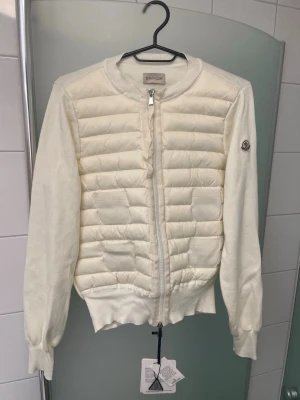 Vit cardiganjacka från Moncler - Snygg vit cardiganjacka från Moncler med quiltad framsida och stickade ärmar. Jackan har dragkedja framtill, ribbade muddar och rund halsringning. Moncler-logga på ärmen och mjukt material som känns lyxigt och bekvämt. NFC funkar 