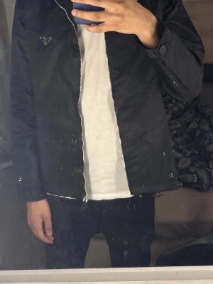 Svart vindjacka med huva - Snygg svart vindjacka med huva och dragkedja framtill. Jackan har en minimalistisk design med en liten logga på bröstet och diskreta fickor. Tillverkad i ett lätt och glansigt material som passar perfekt för blåsiga dagar.