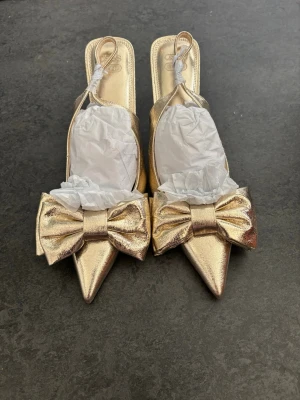 Guldiga pumps med rosett och spetsig tå - Säljer ett par glammiga pumps i metallic guld med spetsig tå och stora dekorativa rosetter framtill. Skorna har öppen häl och smal klack för en elegant look. Perfekta för dig som vill sticka ut och addera lite extra bling till outfiten. Är för stora och helt nya💞