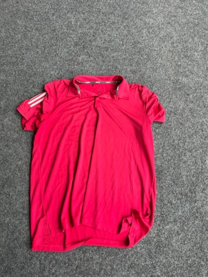 Röd pikétröja från Adidas - Snygg röd pikétröja från Adidas med klassisk krage och tre vita ränder på ena ärmen. Tröjan är kortärmad och har en sportig look, perfekt för träning eller tennis. Tillverkad i ett lätt och funktionellt syntetmaterial.