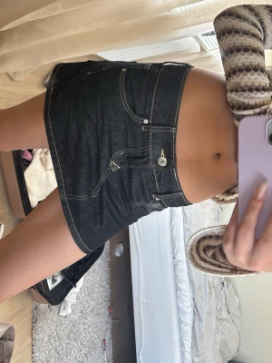 Mörkblå jeanskjol med kontrastsömmar - Säljer en mörkblå kort jeanskjol med tydliga kontrastsömmar och klassiska fickor fram. Kjolen har en snygg, figurnära passform och stängs med knapp och dragkedja. Perfekt för dig som gillar en enkel men trendig look.