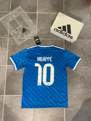Blå Real Madrid fotbollströja Adidas Mbappe - Snygg blå Real Madrid fotbollströja från Adidas med vita detaljer och nummer 10 samt Mbappé på ryggen. Framsidan har klubbmärke och Emirates Fly Better-tryck. Tillverkad i lätt och ventilerande material, perfekt för fotboll eller träning.