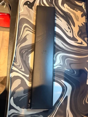 Svart handledsstöd från Razer - Ett svart handledsstöd från Razer, perfekt för gaming eller skrivbordsarbete. Tillverkat i mjukt material med Razer-loggan diskret i mitten. Rektangulär form som passar de flesta tangentbord och ger extra komfort vid långa sessioner.