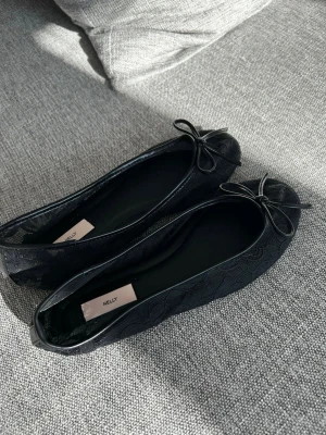 Svarta ballerinaskor med rosett Nelly - Stilrena svarta ballerinaskor från Nelly med söt rosett framtill. Skorna har ett diskret blommönster i tyget och rundad tå. Perfekta för dig som gillar en klassisk look med en twist.