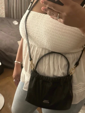 Svart handväska Tommy Hilfiger - Snygg svart handväska från Tommy Hilfiger i mjukt skinnliknande material. Väskan har guldfärgade detaljer, ett kort handtag och en avtagbar axelrem så du kan bära den på olika sätt. Perfekt storlek för det viktigaste och enkel att matcha till olika outfits. Obs! Köpt för 2000kr