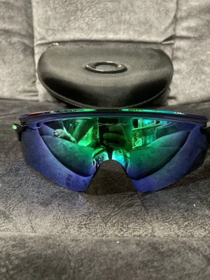 Sportiga solglasögon Oakley.  - Säljer ett par sportiga solglasögon med stora, böjda spegelglas i blå och gröna toner. Glasögonen har en futuristisk look och kommer med ett svart hårt fodral. Perfekta för dig som vill sticka ut och skydda ögonen mot solen.