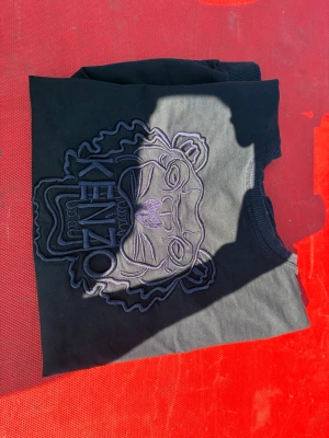 Svart sweatshirt med Kenzo-tryck - Svart sweatshirt från Kenzo med tydlig tigerbrodyr i svart och lila på bröstet. Tröjan har rund halsringning, ribbade muddar och en avslappnad passform. Tröjan är i strl S 