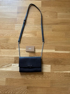 Svart axelväska från BOSS, i läder. - Snygg svart axelväska från BOSS med justerbart axelband i textil och silverfärgade kedjedetaljer. Väskan har stilren design med lock och subtil BOSS-logga framtill. Bredd 21,5 cm Höjd 14,5 cm Djup 3 cm  Inköpspris 2349kr.