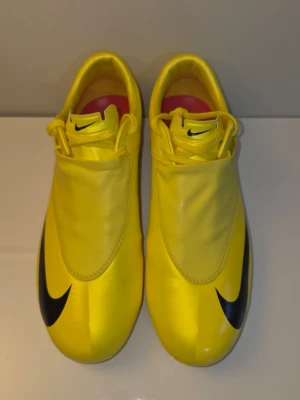 Gula Nike fotbollsskor med svart swoosh - Snygga gula fotbollsskor från Nike med svart swoosh på tå och sidan. Skorna har en slimmad design med täckt snörning och dobbar för bra grepp på planen. Tillverkade i syntetmaterial som ger en lätt och smidig känsla. Perfekta för dig som vill sticka ut på fotbollsplanen.