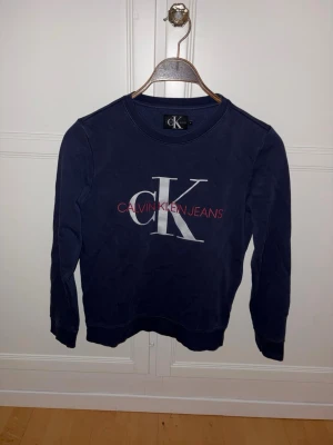 Mörkblå sweatshirt Calvin Klein - Mörkblå sweatshirt från Calvin Klein Jeans. Tröjan har rund halsringning, långa ärmar och ribbade muddar. Perfekt för en avslappnad och stilren look. Storlek 164-176