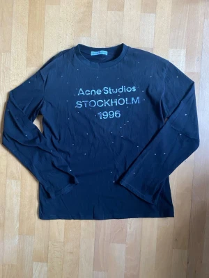 Svart långärmad topp Acne Studios Rhinestone - (Helt Ny) L (passar M) Cool svart långärmad topp från Acne Studios med tryck 'STOCKHOLM 1996' framtill. T-shirten har små silvriga detaljer över hela framsidan och är gjord i mjuk bomull. Perfekt till jeans eller kjol för en trendig look. (Rhinestone)