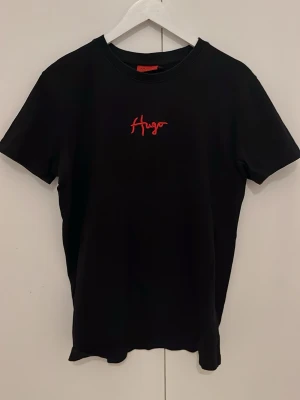 Svart t-shirt från Hugo med röd logga - Snygg svart t-shirt från Hugo med röd logga broderad på bröstet. Klassisk passform och rund halsringning. Perfekt till jeans eller shorts för en clean och stilren look.