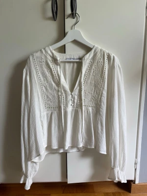Vit blus med spetsdetaljer - Säljer en vit boho-inspirerad blus med broderade spetsdetaljer framtill och knappar vid urringningen. Blusen har lång ärm med knyt vid ärmslut och är gjord i ett lätt, luftigt tyg som ger en avslappnad vibe. Välj gärna instabox😊