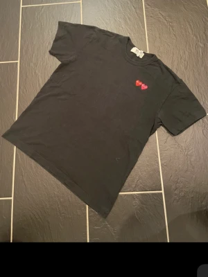 Svart Comme des Garçons PLAY t-shirt - Svart t-shirt från Comme des Garçons PLAY med två broderade hjärtan i rött och vinrött på bröstet. Klassisk rund halsringning och korta ärmar. Perfekt för dig som gillar stilrena plagg med en twist.