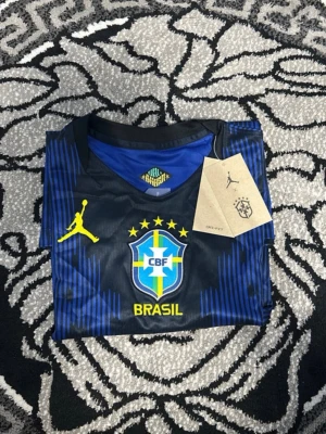 Blå Brasilien fotbollströja Jordan - Snygg blå och svart Brasilien fotbollströja från Jordan med gula detaljer och CBF-logga på bröstet. Tröjan har korta ärmar, rund hals och är tillverkad i ett lätt, ventilerande material som passar perfekt till träning eller match.