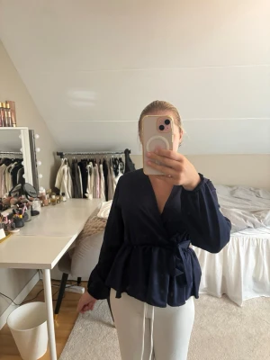 Mörkblå omlottblus med knytband - Snygg mörkblå omlottblus med knytband i midjan och v-ringning. Blusen har långa ärmar och en lätt volang nedtill som ger en feminin siluett. Perfekt att styla med både jeans och kjol för en trendig look.