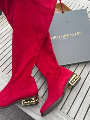 Röda overknee stövlar i mocka (36) - Snygga röda overknee stövlar från Pedro Miralles i mjuk mocka. Stövlarna har låg klack med guldfärgade nitar som detalj och snörning upptill för perfekt passform. Perfekta för att sticka ut och ge din outfit en färgklick.