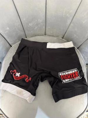 Svarta MMA-shorts Fighter Gear - Säljer ett par svarta MMA-shorts med vita detaljer och tryck. På ena benet står det 'Fighter Gear' i rött och vitt, och på andra benet finns ett rött och vitt märke med texten 'Standing Tall'. Perfekta för kampsportsträning eller grappling.