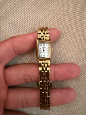 Mockberg Klocka - Stilren klocka med rektangulärt vitt ur och romerska siffror. Armbandet är guldfärgat i metall med klassisk länkdesign. Perfekt accessoar för dig som gillar tidlös och elegant stil.