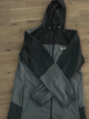 Grå och svart vindjacka Under Armour - Snygg vindjacka från Under Armour i grått och svart med huva och dragkedja framtill. Jackan har elastiska muddar vid ärmsluten och logga på bröstet samt text på ryggen. Perfekt för blåsiga dagar och sportiga outfits.