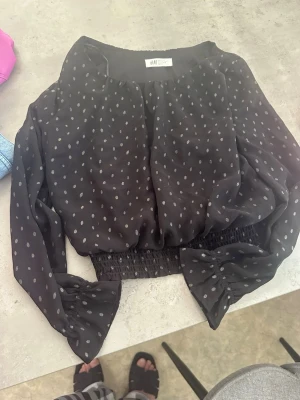 Svart prickig blus från H&M. - Svart blus från H&M med silvriga prickar och lång ärm. Blusen har resår i midjan och vid ärmsluten för en ballongig look. Tillverkad i ett tunt, luftigt material som känns skönt mot huden. Perfekt för dig som gillar trendiga detaljer och en avslappnad men snygg stil.