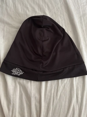 Svart tunn mössa med brodyr - Svart, slimmad mössa i stretchigt material med diskret vit brodyr på kanten. Perfekt för dig som gillar en enkel och stilren look. Mössan är lätt och passar bra under jackhuvan eller hjälmen.
