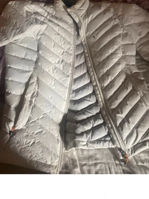 Vit dunjacka från Peak Performance - Snygg vit dunjacka från Peak Performance med dragkedja och två sidofickor. Jackan har ett stilrent, quiltat mönster och lätt glansigt material. Perfekt för kalla vinterdagar och riktigt skön att bära. Klassisk logga på bröstet.