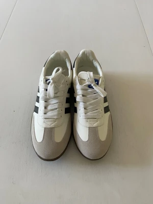 Vita sneakers med svarta ränder - Snygga vita sneakers med klassiska svarta ränder på sidorna och beige mockadetaljer framtill. Skorna har vit ovandel, svart hälparti och bruna yttersulor. Perfekta för en sportig och stilren look. Lågt skaft och snörning framtill.