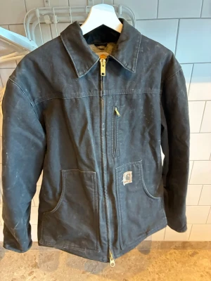 Carhartt Jacka- L/XL - Klassisk svart workwear jacka i robust bomullsmaterial med guldfärgad dragkedja och stor krage. Jackan har två stora framfickor, en bröstficka med dragkedja och en liten logotyp på vänster sida. Perfekt för dig som gillar en enkel och tidlös stil. 