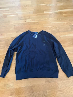 Mörkblå stickad tröja Polo Ralph Lauren - Mörkblå stickad tröja från Polo Ralph Lauren med rund halsringning och klassisk logga broderad på bröstet. Tröjan har långa ärmar och ribbade muddar vid ärmslut och nederkant. Perfekt för en stilren och enkel look.