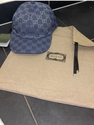 Blå keps från Gucsi i denim - Säljer en snygg blå keps från Gusci i denim med det klassiska GG-mönstret över hela kepsen. Böjd skärm och justerbar baksida. Perfekt för dig som vill ha en trendig accessoar med lyxig känsla och streetwear-vibe.