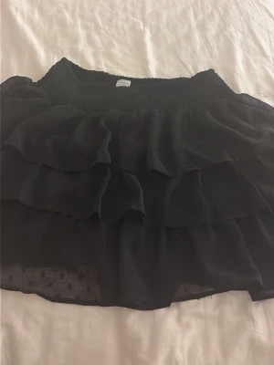 Svart volangkjol med resårmidja - Säljer en svart kortkjol med flera lager volanger och bred resår i midjan. Kjolen har en luftig och flowy känsla, perfekt för dig som gillar en feminin och trendig stil. Materialet känns lätt och skirt.