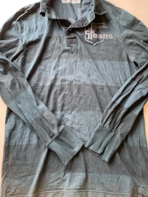 Randig långärmad henley från Calvin Klein - Säljer (priset kan diskuteras) en långärmad henley från Calvin Klein Jeans i blå och grå bredrandig design. Skjortan har krage, knappar vid halsen och broderad logga på bröstet. Perfekt för dig som gillar avslappnad men snygg stil. Den ser grå ut men är blågrön i verkligen 