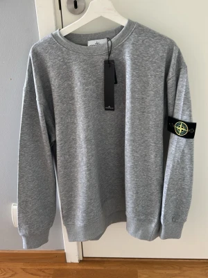 Grå sweatshirt från Stone Island - Snygg grå sweatshirt från Stone Island med klassisk logotyp-patch på ärmen. Tröjan har rund halsringning, ribbade muddar och en relaxed passform. Perfekt för en stilren och avslappnad look.