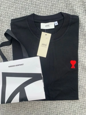 Svart t-shirt från Ami Paris - Svart t-shirt från Ami Paris med klassisk passform och rund hals. T-shirten har en liten röd broderad logga i form av ett hjärta på bröstet. Tillverkad i mjuk bomull och perfekt för en stilren look.