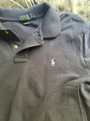 Mörkblå pikétröja från Polo Ralph Lauren - Klassisk mörkblå pikétröja från Polo Ralph Lauren med broderad logga på bröstet. Tröjan har krage, knappar framtill och är tillverkad i bomull. Perfekt för en stilren och avslappnad look. Äkta, kvitto finns ej med eftersom denna är köpt från en second hand butik i newyork City i USA. Storlek L. Inga skador eller liknande.